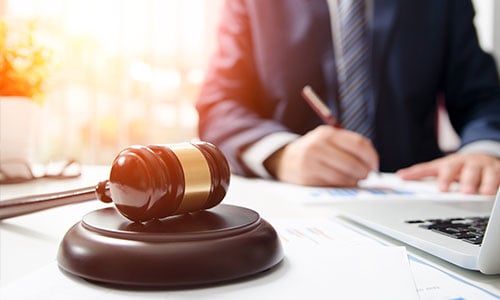 Ein Richterhammer im Sonnenschein. Im Hintergrund: Mann im Anzug